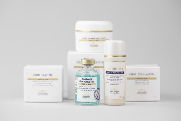 Dwie buteleczki kosmetyczne na pierwszym planie — szklana fiolka z niebieskim serum oznaczona „LIPOSMOSE Sérum Authentique” i plastikowa butelka „Lotion P50” — przed kilkoma białymi pudełkami kremów na jasnym neutralnym tle.