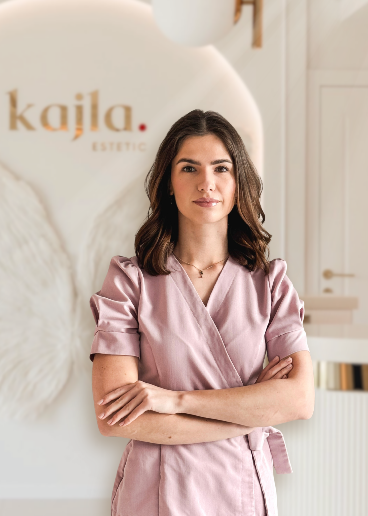 Kobieta o ciemnych, lekko falowanych włosach stoi z założonymi rękami w pastelowo-różowym uniformie kosmetycznym przed jasnym, eleganckim wnętrzem gabinetu z widocznym logo „kajla estetic”.