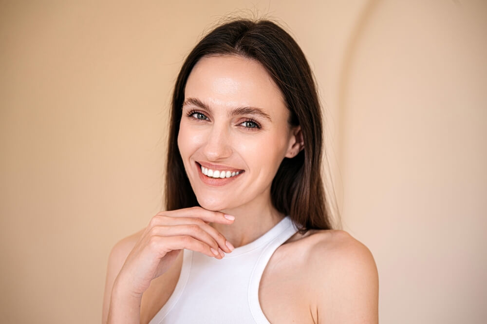 Uśmiechnięta kobieta z długimi ciemnymi włosami w białym topie, opiera dłoń na podbródku i patrzy w obiektyw na jednolitym beżowym tle.